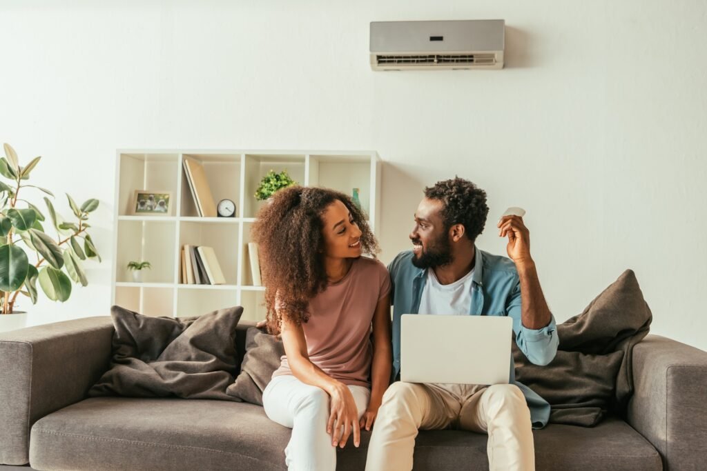 handsome african american man using laptop and holding air conditioner remote controller while